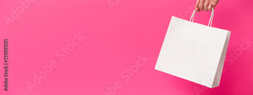 Female hand holding white blank shopping bag on pink background. White craft blank paper bag in hand. Discount, shopping and ecology concept. Black Friday sale banner