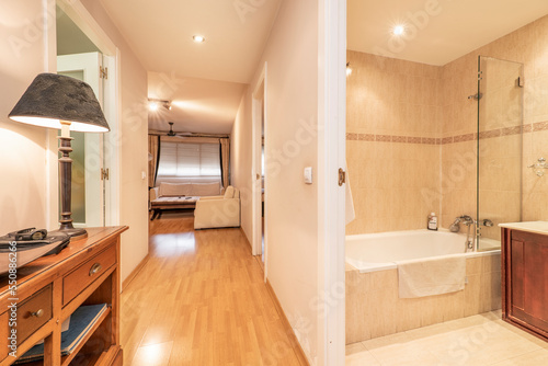 Distributor corridor of the house with floating oak flooring  access to a bathroom with a tub and in the background a living room with white fabric armchairs