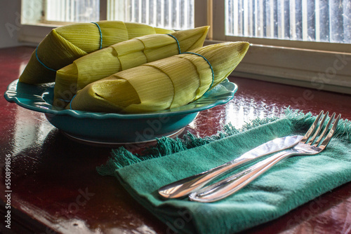 Três pamonhas embrulhadas em palha verde, dentro de um prato, sobre uma superfície reluzente de frente à uma janela de vidro. photo