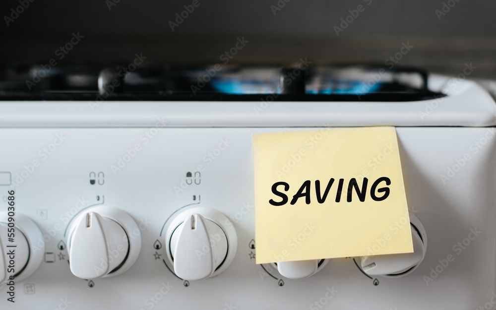 Home energy saving gas concept. Text on sticker Saving pasted on panel of gas stove with burning burner indoors, close-up Home energy saving gas concept. Text on sticker Saving pasted on panel of gas stove with burning burner indoors, close-up