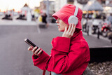 Stylish woman in magenta color jacket and bucket hat wearing wireless headphones on her head and setting phone for listening music. Urban city street fashion. Color of the 2023 year. Selective focus.
