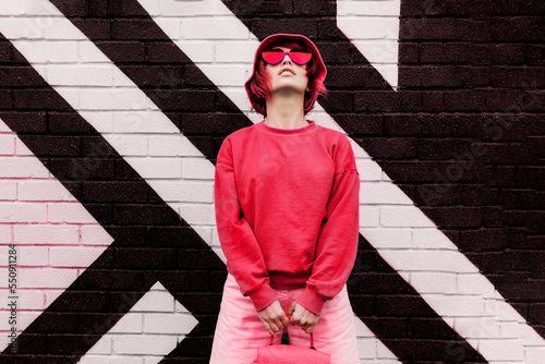 Hipster fashion young woman in trendy magenta color sweatshirt and sunglasses and bucket hat posing on the painted brick wall background. Color of the 2023 year. Urban city street fashion.