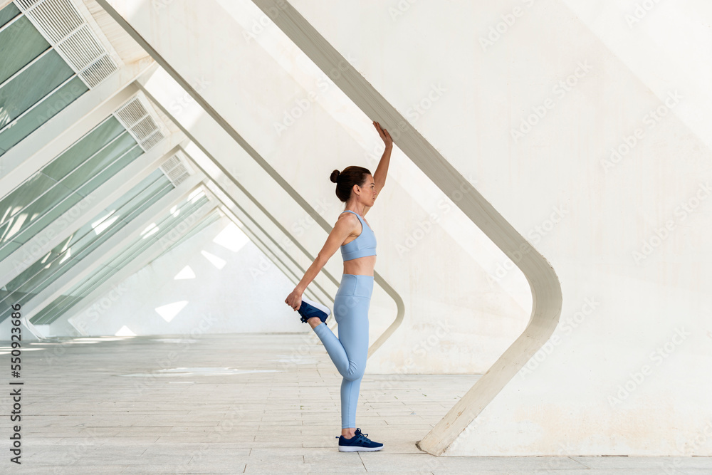 Sporty woman doing a leg stretch, warm up exercise. Modern urban background.