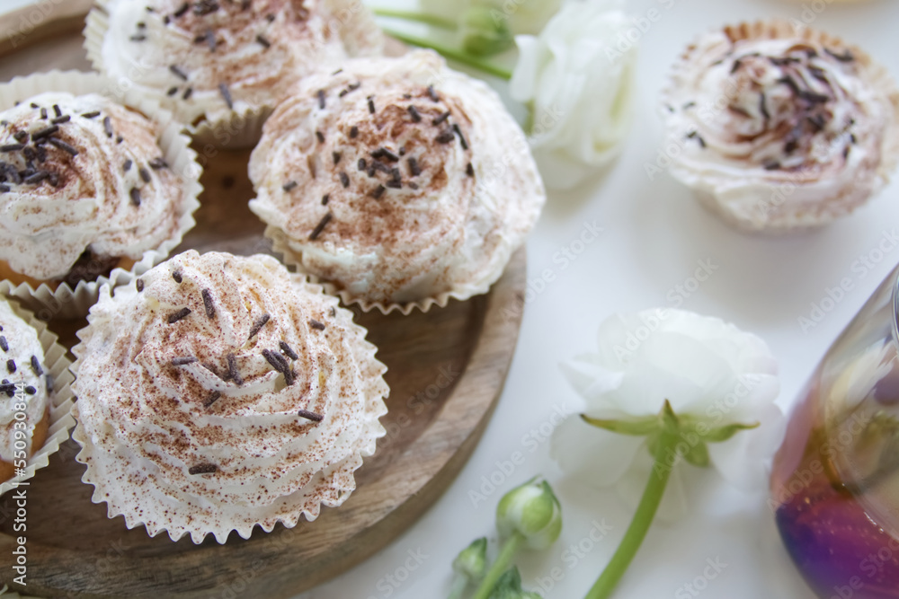 Muffins with cream and chocolate sprinkles on a wooden stand near white ranunculus and a cup of tea Muffins with cream and chocolate sprinkles on a wooden stand near white ranunculus and a cup of tea