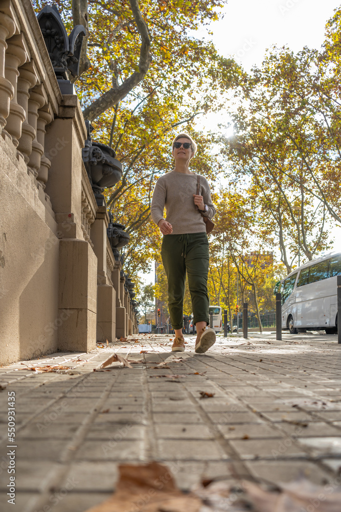 Blond woman with short hair in sweater and backpack walking through autumn city in Barcelona.