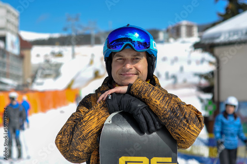 A happy and contented snowboarder stands with a snowboard near the ski lifts. Ski resort in the mountains.