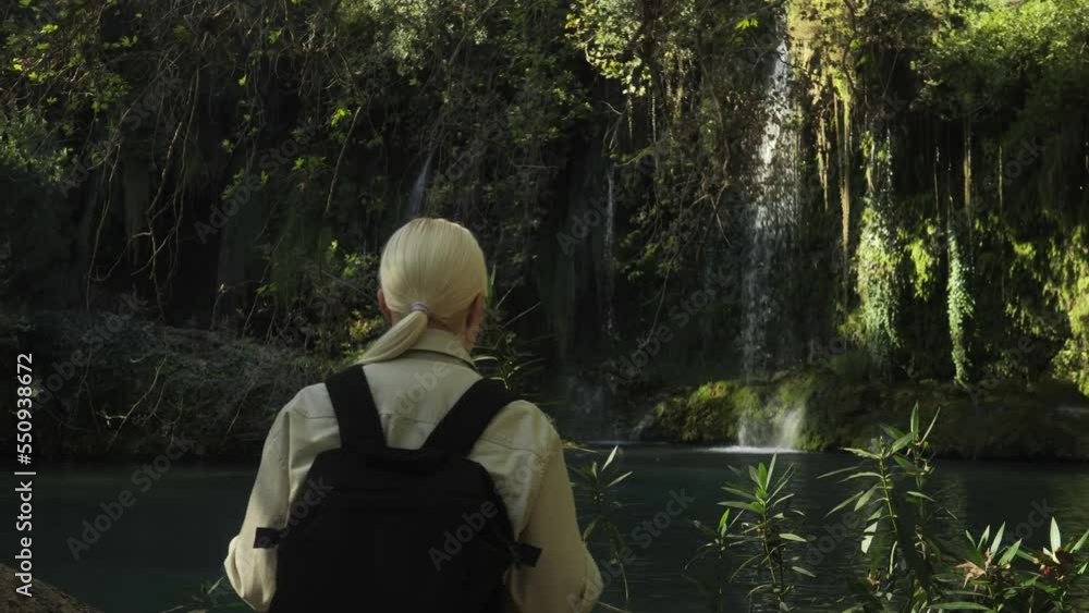 A pretty young blonde tourist woman comes out of the forest to a beautiful forest waterfall. The concept of opening a new one, the beauty of wild nature. High quality 4k footage A pretty young blonde tourist woman comes out of the forest to a beautiful forest waterfall. The concept of opening a new one, the beauty of wild nature. High quality 4k footage