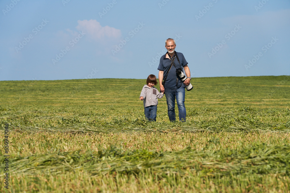Grandfather and granddaughter spending time together. An adult man and a little girl holding hands walk across the field. The man has a camera. They are happy to be together.