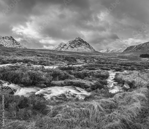 Black and white Majestic Winter landscape image of River Etive in foreground with iconic snowcapped Stob Dearg Buachaille Etive Mor mountain in the background
