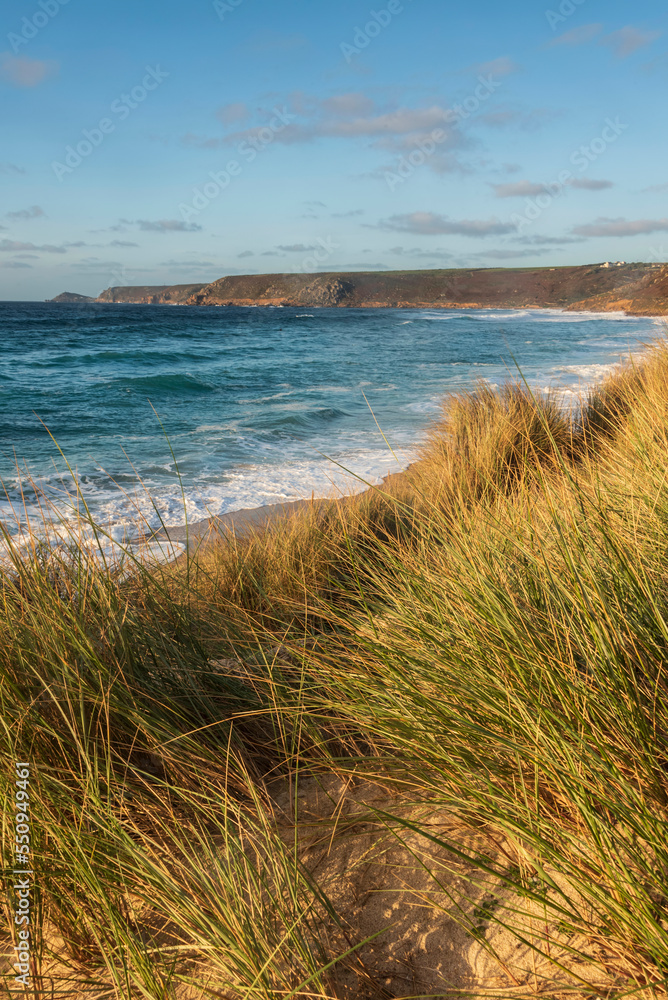 Stunning landscape image of Sennen Cove in Cornwall during sunset viewed from grassy sand dunes with dramatic sky and long exposure sea motion