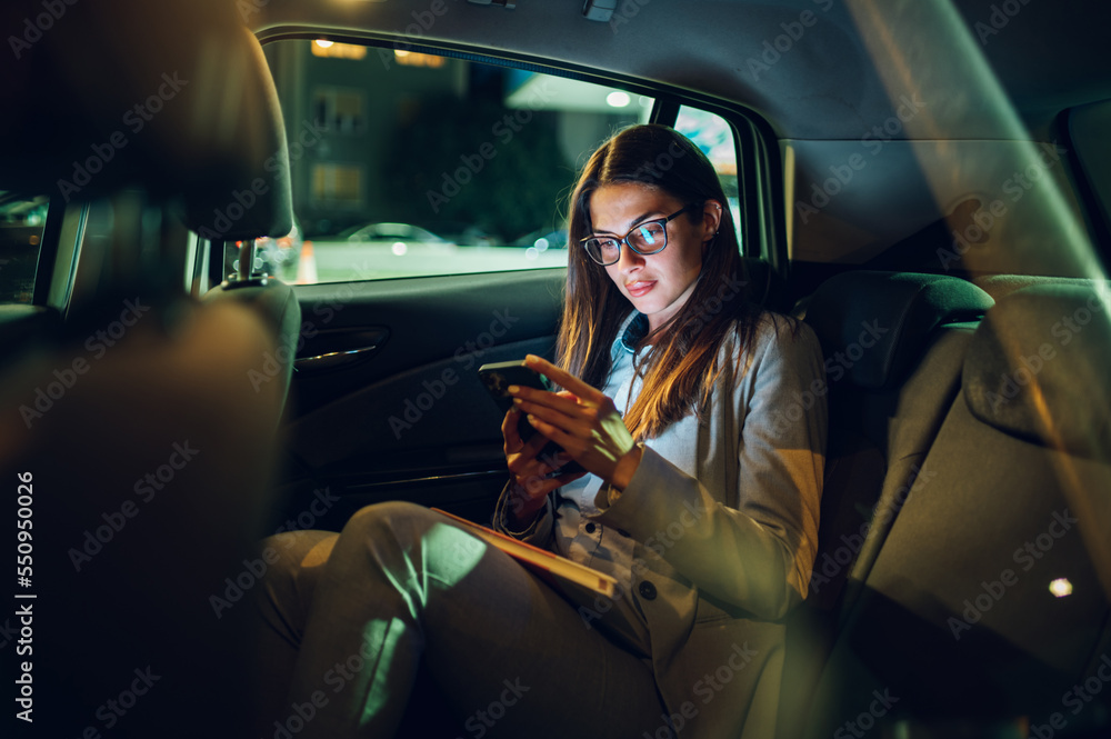 Business woman using smartphone while sitting in a backseat of a car at night - obrazy, fototapety, plakaty 