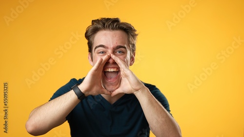 Portrait of happy young man 20s smiling scream and shout calling inviting with hands at mouth say hey you isolated on solid yellow background. photo