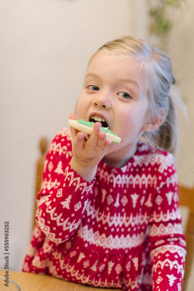 Little girl in a red Christmas sweater tasting a cookie she just decorated Little girl in a red Christmas sweater tasting a cookie she just decorated