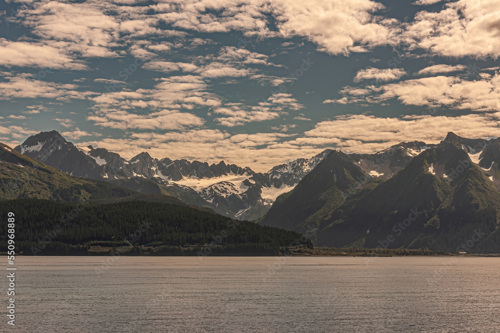 Seward, Alaska, USA - July 22, 2011: Wide landscape of Resurrection bay shoreline with snow covered mountain range and green forested flanks under blue cloudscape