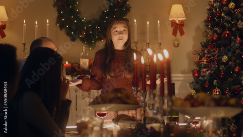 Young asian girl tells joke, poem or plays game with large multi cultural family. They celebrating Christmas. Served table with dishes and candles. Warm atmosphere of family Christmas dinner at home.
