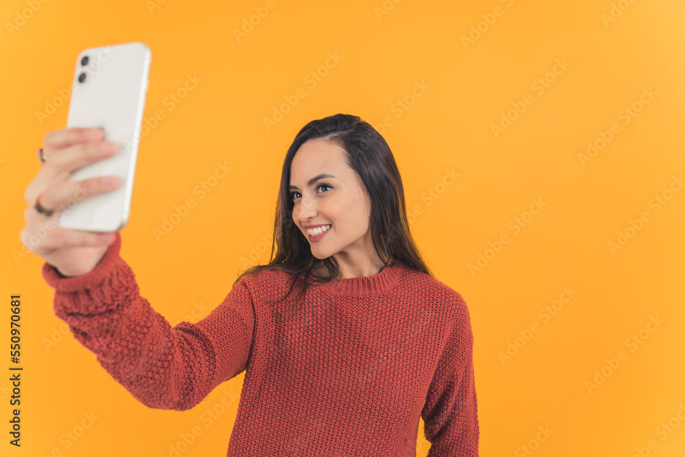 Latin american young adult woman with dark long hair wearing red sweater holding smartphone taking selfie smiling. Yellow background horizontal studio shot. High quality photo Latin american young adult woman with dark long hair wearing red sweater holding smartphone taking selfie smiling. Yellow background horizontal studio shot. High quality photo