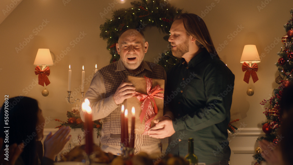 Man giving gift to his senior father. Happy family celebrating Christmas or New Year 2023. Served holiday table with candles. Warm atmosphere of family Christmas dinner at home.