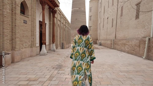 4K. Overall plan. Tourism in Uzbekistan. An oriental girl in a traditional Uzbek robe walks through the historical center of Bukhara. Back view.