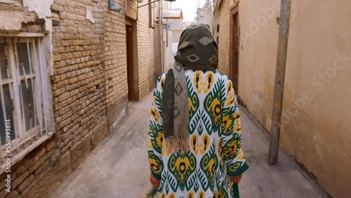 A girl in an Uzbek traditional robe walks through the old city of Bukhara. Medium plan. 4K. Back view. An oriental girl walks among the residential buildings of the old city. Tourism in Uzbekistan.