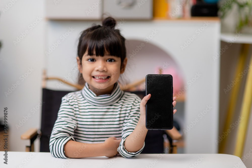 Cute little girl uses smartphone while sitting at the sofa in the living room. Child surfing the internet on mobile phone, browses through internet and watches cartoons online at home Cute little girl uses smartphone while sitting at the sofa in the living room. Child surfing the internet on mobile phone, browses through internet and watches cartoons online at home