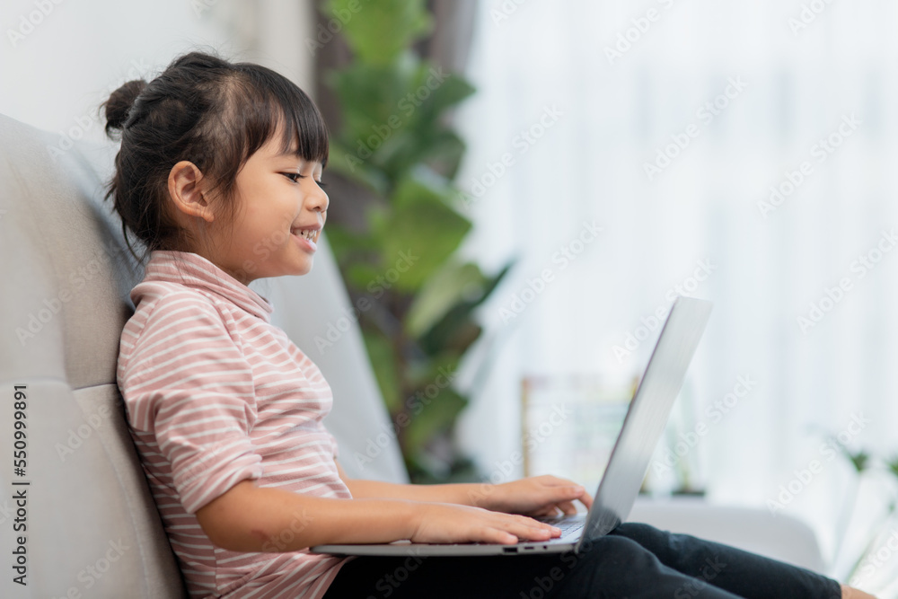 Asian Little school kid girl use laptop computer sitting on sofa alone at home. Child learning reading online social media content, play education lessons game chatting with friends. Asian Little school kid girl use laptop computer sitting on sofa alone at home. Child learning reading online social media content, play education lessons game chatting with friends.