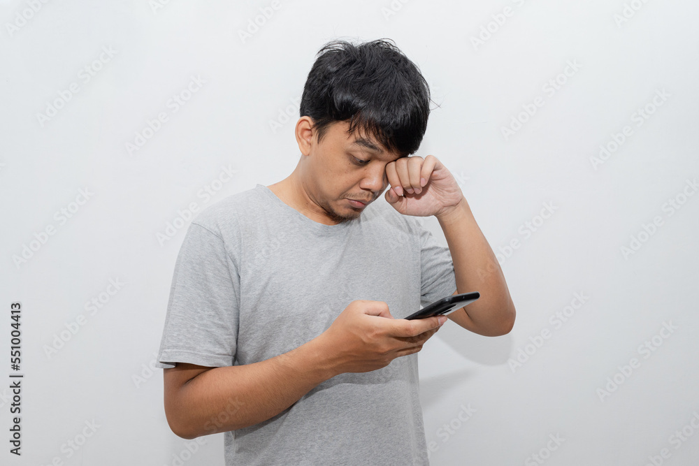 Asian young man rubs his eye and looks at smartphone after wake up from sleep with white background Asian young man rubs his eye and looks at smartphone after wake up from sleep with white background