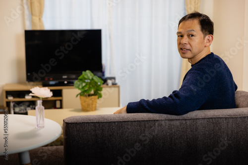 A middle-aged Asian man relaxes on a charcoal gray couch in a living room.