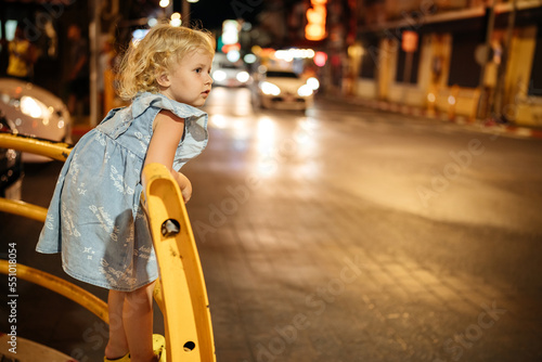 A 4-5 year old child stands at a pedestrian crossing and waits for a traffic light in the evening with car headlights.