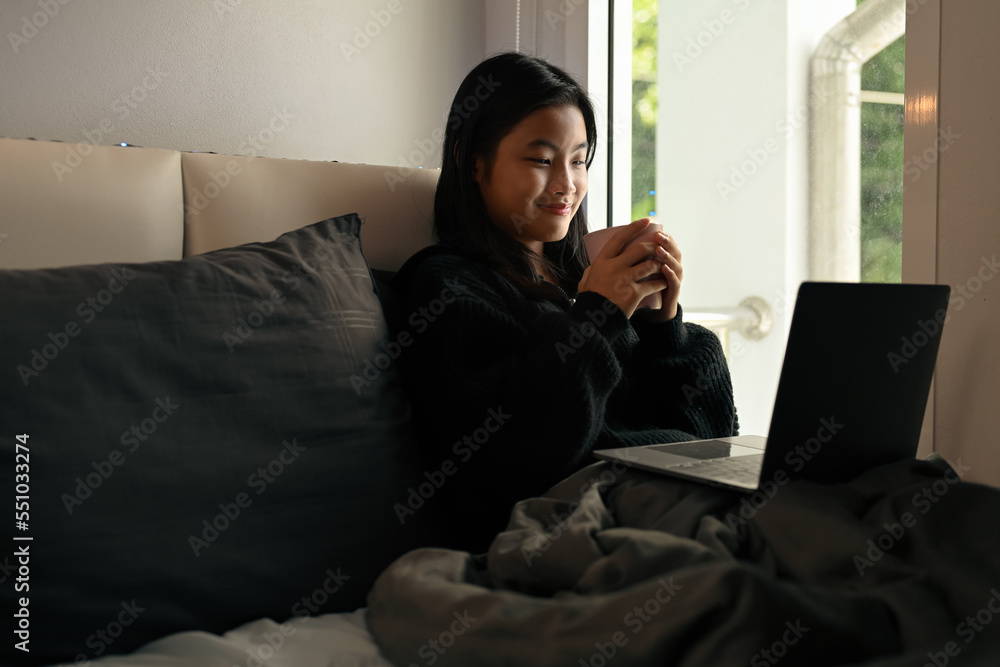 Smiling asian girl drinking hot chocolate and using laptop on bed, relaxing on weekend morning Smiling asian girl drinking hot chocolate and using laptop on bed, relaxing on weekend morning