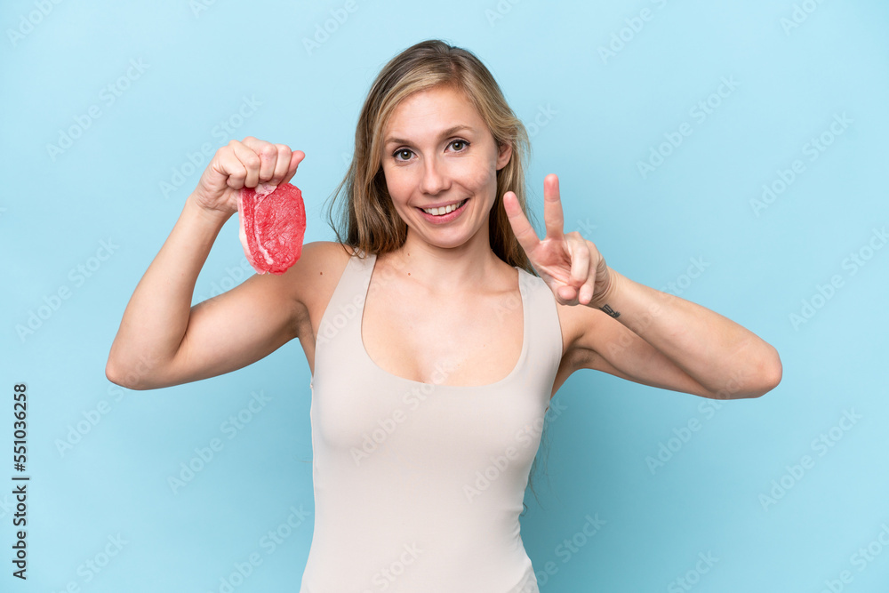 Young blonde woman holding a piece of meat isolated on blue background smiling and showing victory sign Young blonde woman holding a piece of meat isolated on blue background smiling and showing victory sign