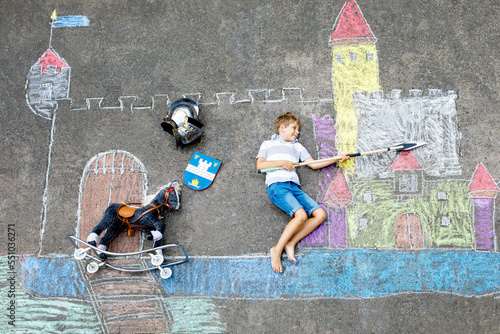 Little active kid boy drawing knight castle and fortress with colorful chalks on asphalt. Happy child in helmet and with spear having fun with playing knight game and painting
