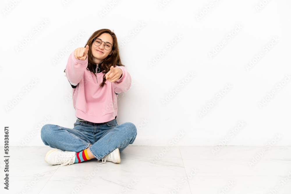Young caucasian woman sitting on the floor isolated on white background points finger at you while smiling