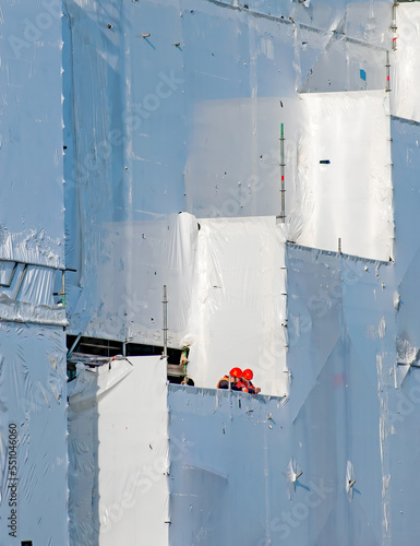 Two construction workers look out from an unusual landscape of white plastic sheeting. The stark white sheeting follows a big stepped shape form with just the two workers in bright orange centrally. 