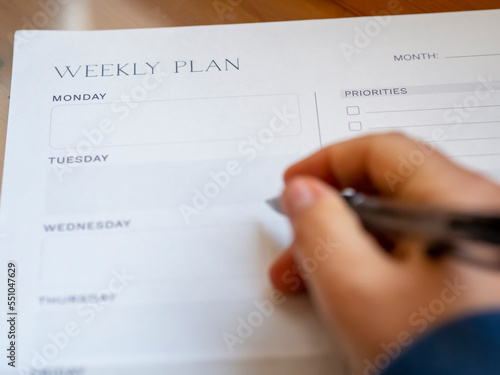 Woman's hand-holding pen and weekly planning on a blank daily schedule. Week planner for organization and goal priorities. Writing down tasks and schedule reminders on office desk.