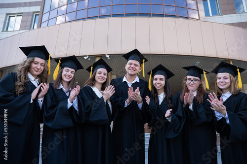 Seven graduates in robes stand in a row and clap outdoors.