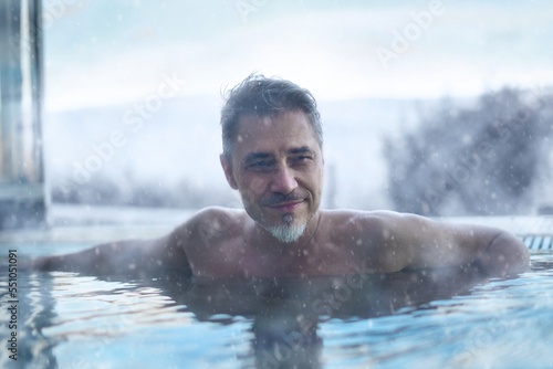 Happy older man enjoying hot thermal water in cold winter weather in snow. Relaxing bath in outdoor whirl pool in wellness spa. Happy mid adult, middle aged guy smiling. © nyul