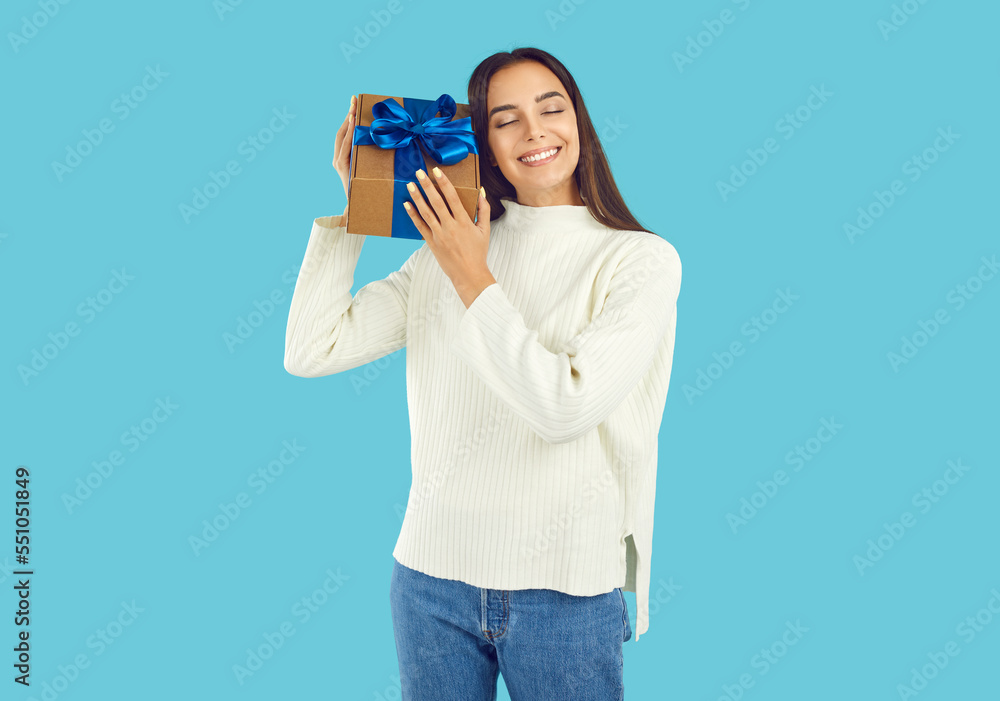 Portrait of happy beautiful young woman in warm white winter sweater standing on blue background, holding gift box with Christmas present tied with blue bow and smiling with eyes closed Portrait of happy beautiful young woman in warm white winter sweater standing on blue background, holding gift box with Christmas present tied with blue bow and smiling with eyes closed