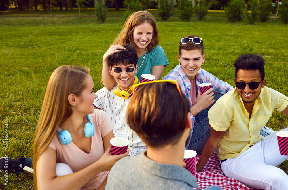 Young positive group of friends men and women vacationers on picnic drinking drinks from paper cups having fun after college or university sits on meadow in park. Student leisure lifestyle Young positive group of friends men and women vacationers on picnic drinking drinks from paper cups having fun after college or university sits on meadow in park. Student leisure lifestyle