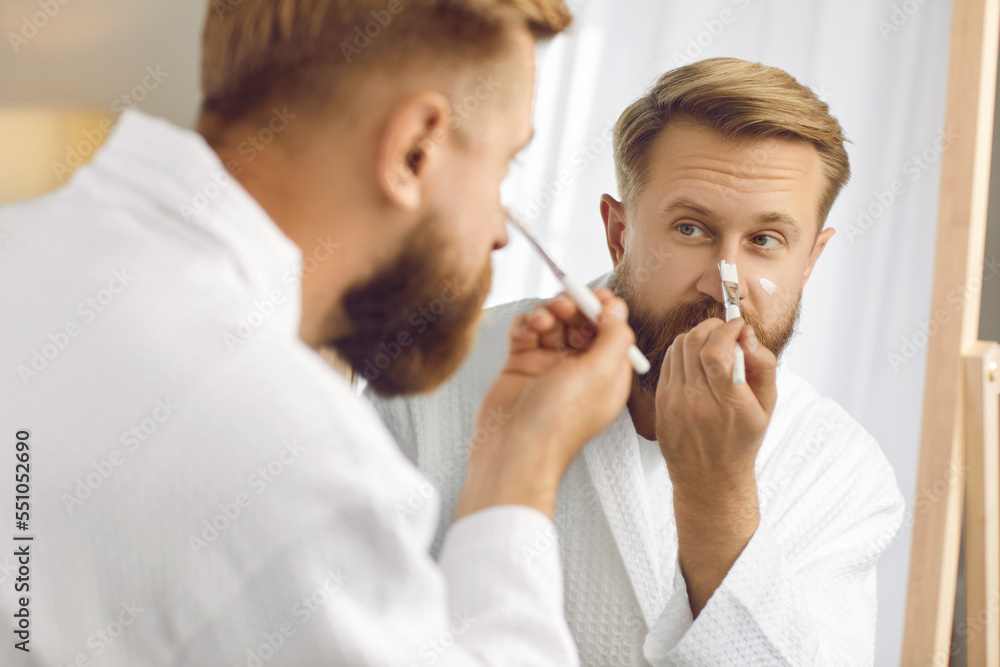 Cosmetics for men. Handsome bearded man with brush applies moisturizing mask on his face during home beauty treatments. Close up of caucasian millennial man in bathrobe looking in mirror. Cosmetics for men. Handsome bearded man with brush applies moisturizing mask on his face during home beauty treatments. Close up of caucasian millennial man in bathrobe looking in mirror.
