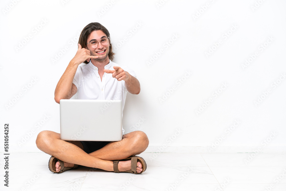 Young handsome man with a laptop sitting on the floor isolated on white background making phone gesture and pointing front Young handsome man with a laptop sitting on the floor isolated on white background making phone gesture and pointing front