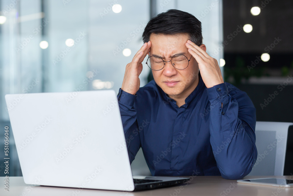 Asian businessman boss sick has severe headache, man in glasses sitting at desk using laptop at work inside office, migraine office worker investor overtired. Asian businessman boss sick has severe headache, man in glasses sitting at desk using laptop at work inside office, migraine office worker investor overtired.