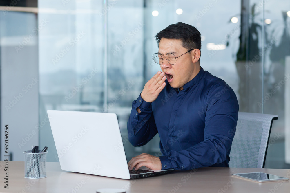 Tired worker at work yawning, asian man in shirt and glasses working inside office using laptop, man sleepless at work. Tired worker at work yawning, asian man in shirt and glasses working inside office using laptop, man sleepless at work.