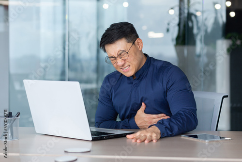 Mature asian businessman sick in office, man having heart attack, senior boss holding hands on chest heartache, boss working inside office using laptop at work.