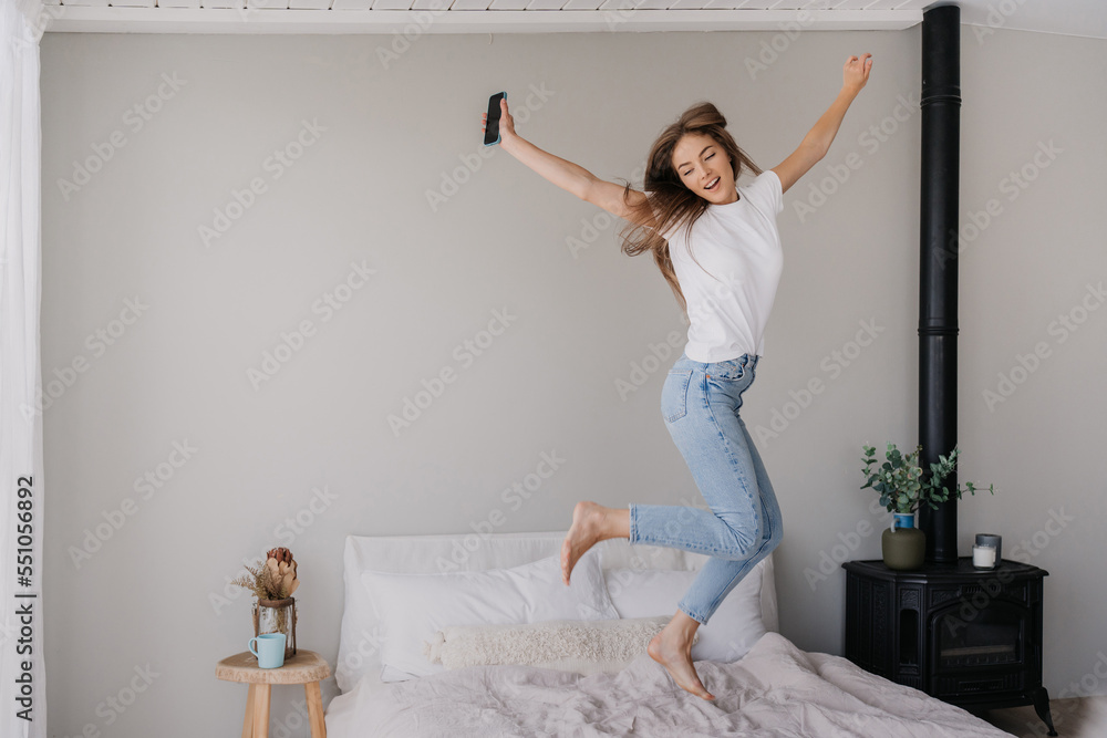Jumping hi young adult Italian woman in white t-shirt and blue denim jeans holds phone happy jumps on bed at home. Mockup with copy space with celebrating Caucasian girl. Happiness and youth. Jumping hi young adult Italian woman in white t-shirt and blue denim jeans holds phone happy jumps on bed at home. Mockup with copy space with celebrating Caucasian girl. Happiness and youth.