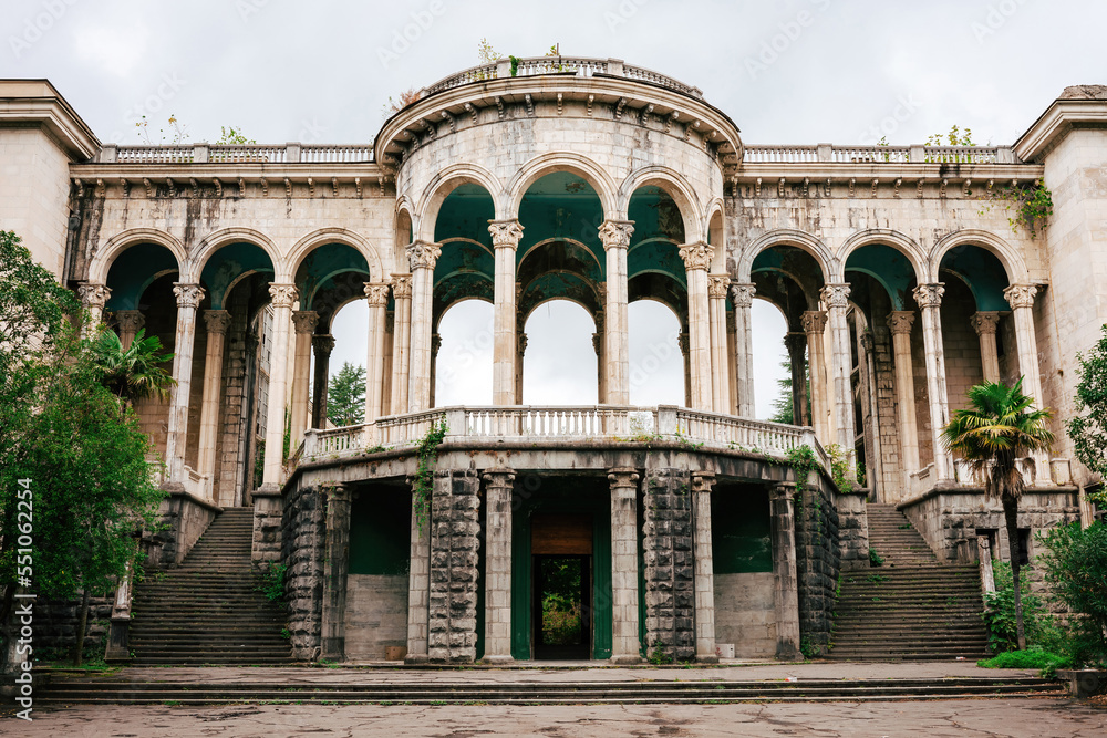 Facade of a decaying palace or hotel with tall marble columns and arches. The abandoned building is surrounded by bushes against a gray sky