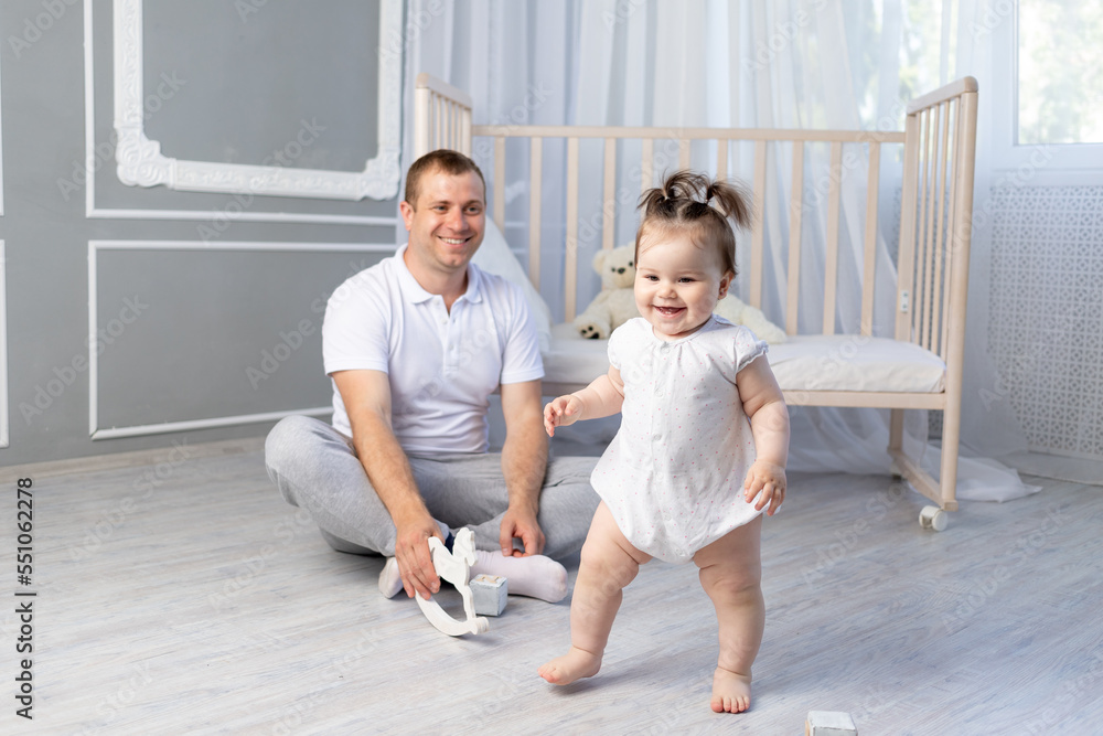 baby girl takes her first steps, dad teaches baby to walk in the nursery, happy fatherhood baby girl takes her first steps, dad teaches baby to walk in the nursery, happy fatherhood