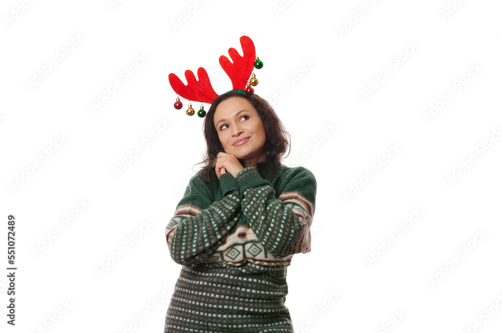 Charming Latin American pretty woman in green Xmas patterned sweater and deer antler hoop, sweetly smiling, dreamily looking away at a copy space on white background. Christmas and New Year concept Charming Latin American pretty woman in green Xmas patterned sweater and deer antler hoop, sweetly smiling, dreamily looking away at a copy space on white background. Christmas and New Year concept