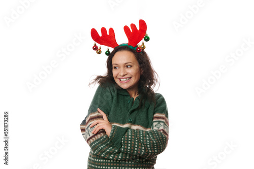 Multi-ethnic joyful attractive woman, wearing deer antler hoop and warm green sweater with Christmas pattern, hugging herself, laughing, looking aside at copy advertising space on white background