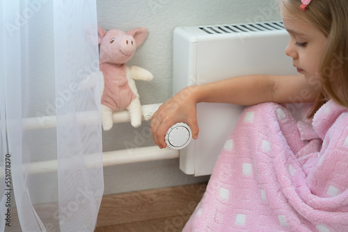 Close up of little girl sit in plaid near heating radiator and adjust thermostat regulator. Central heating system. Heat saving.