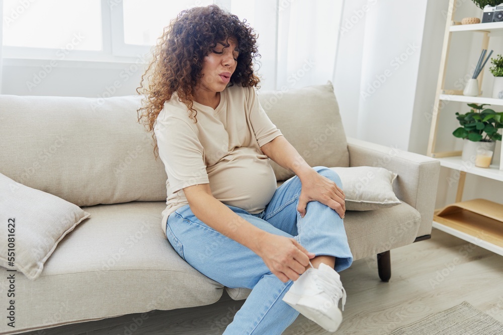 Pregnant woman sitting on sofa at home and putting on pregnancy-friendly shoes with problem due to belly, headache Pregnant woman sitting on sofa at home and putting on pregnancy-friendly shoes with problem due to belly, headache